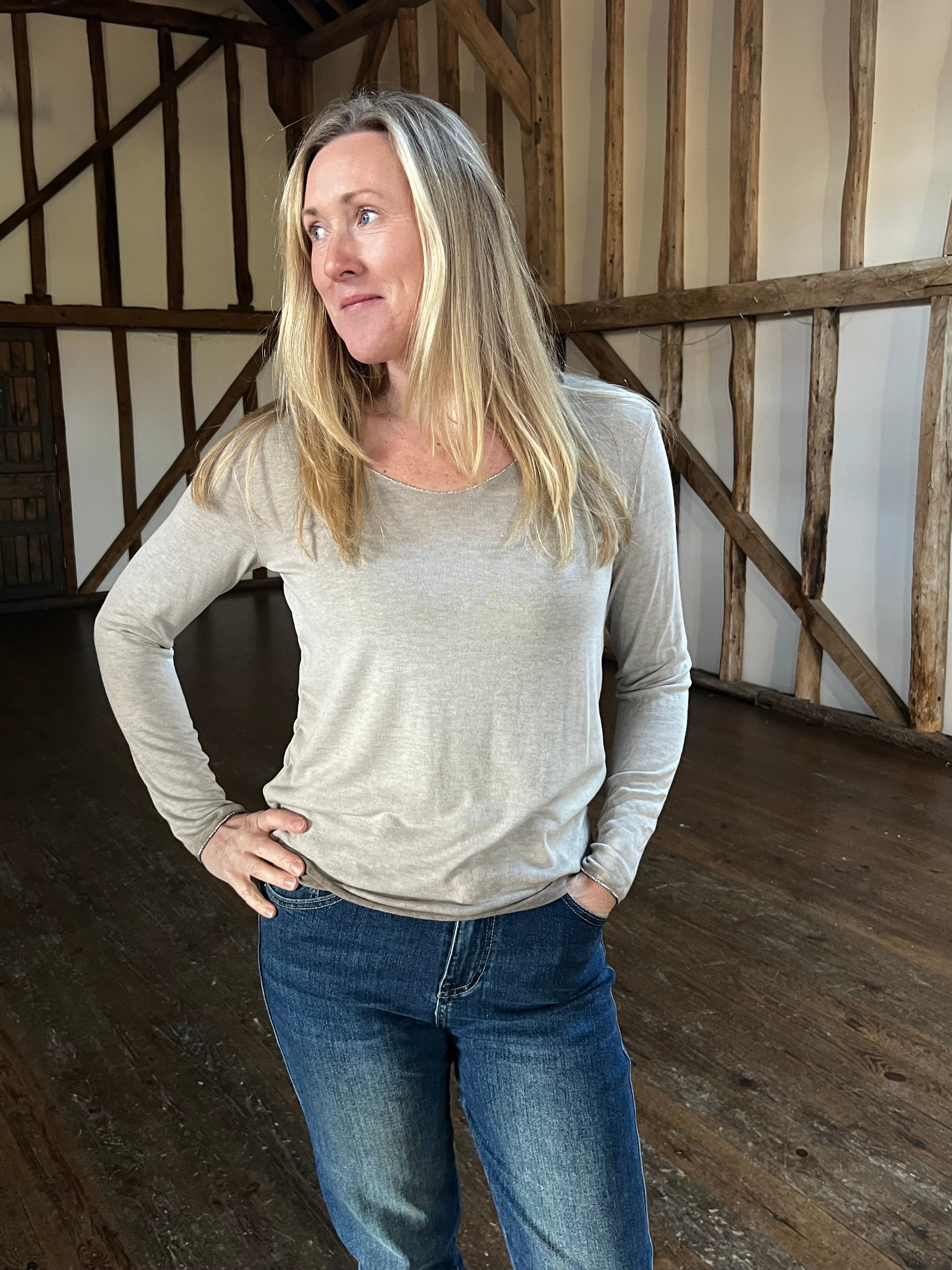 A woman standing in a room with wooden walls, wearing a long-sleeved, striped, sparkly top and blue jeans.