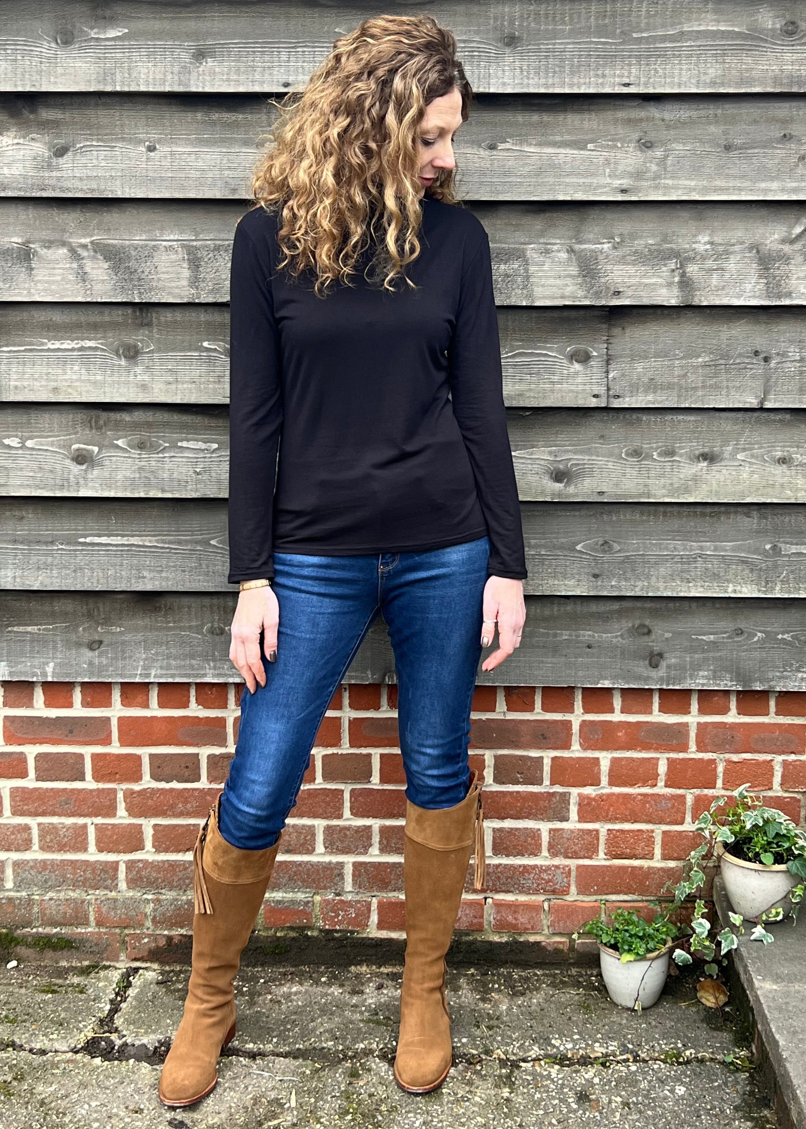 Woman wearing a black long-sleeve top and blue jeans standing against a wooden wall.