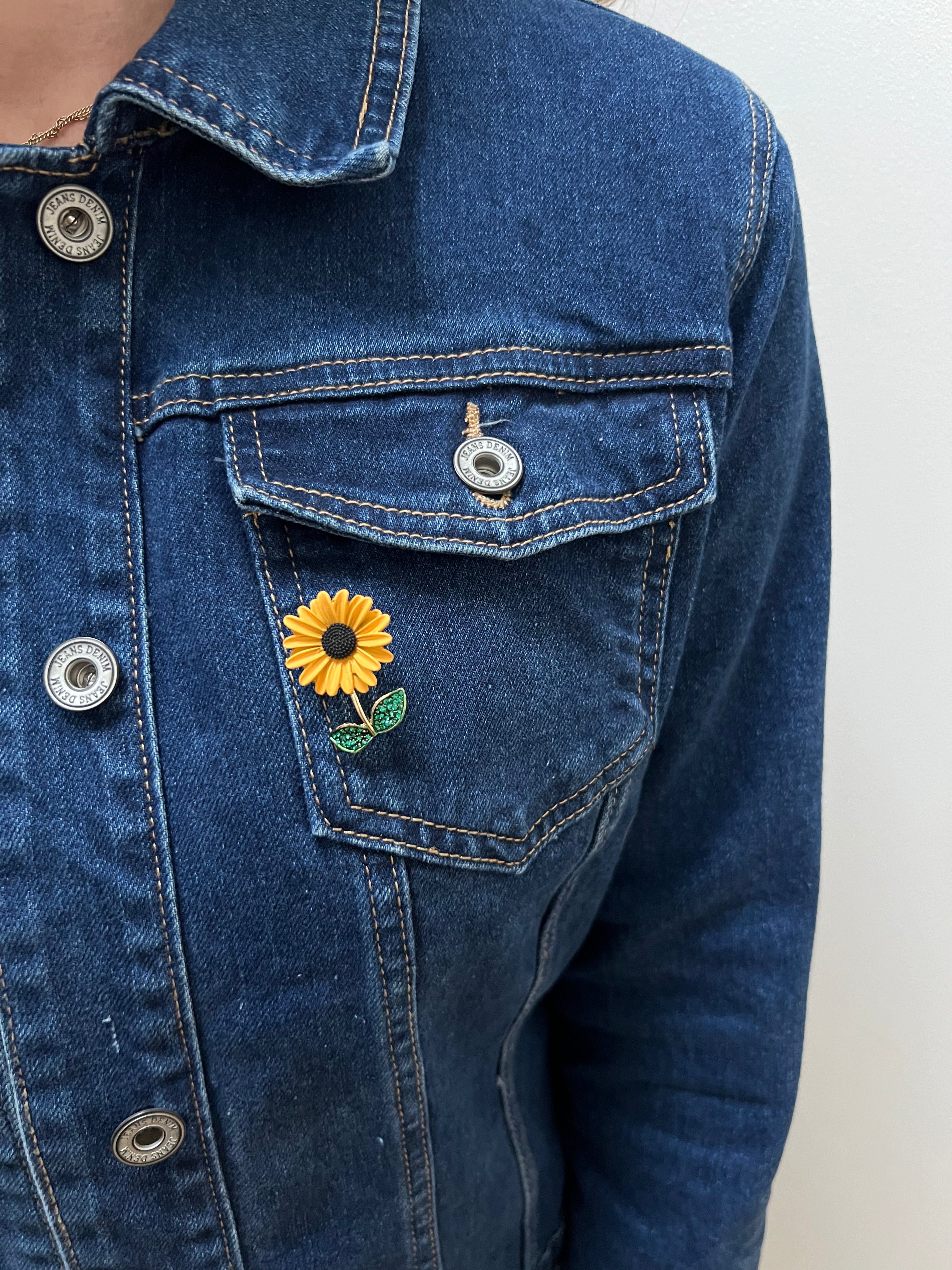 A person wearing a blue denim jacket with a sunflower brooch on the lapel.