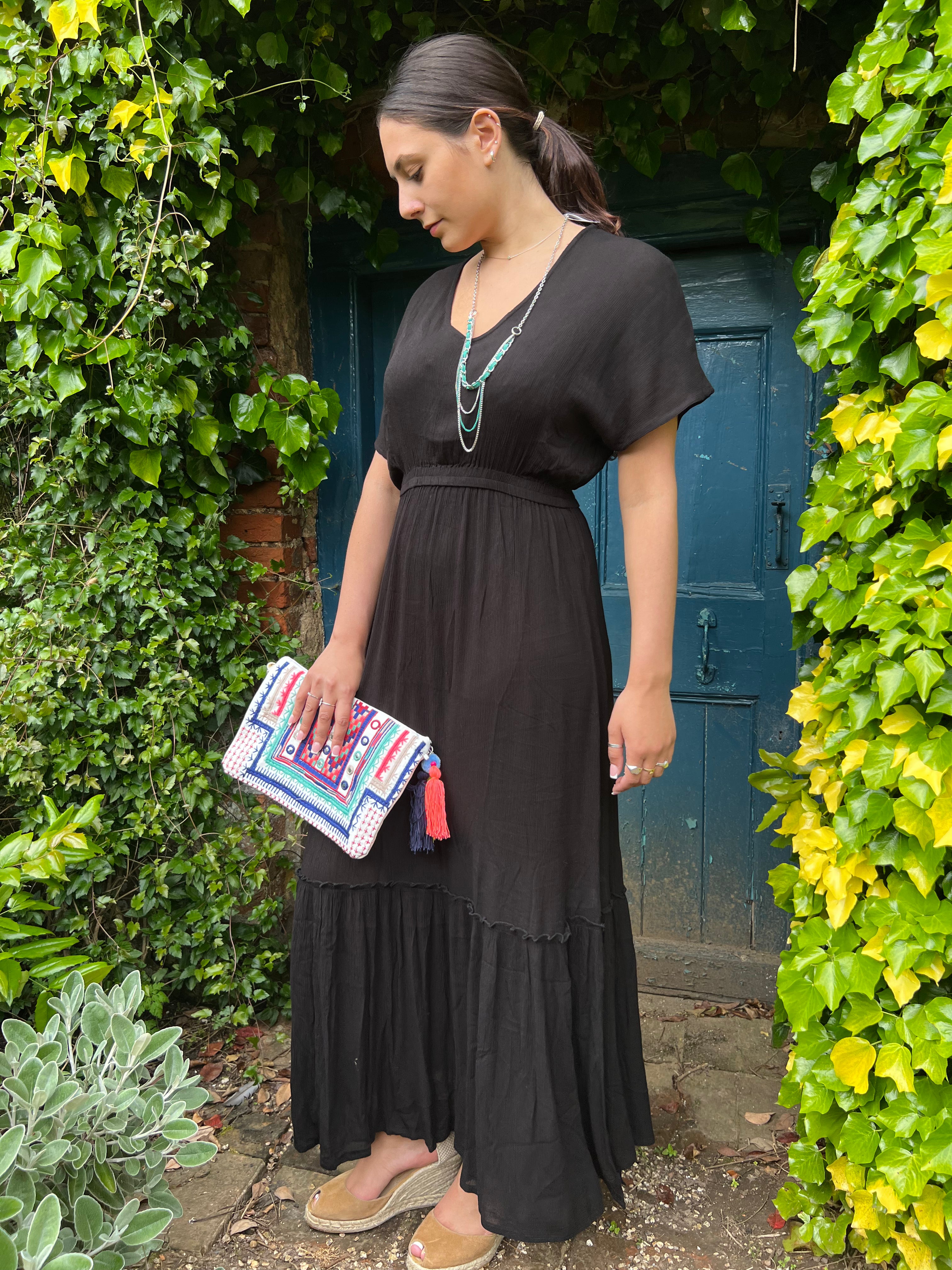 Person standing wearing a talia maxi dress in black by a door outside and trees.