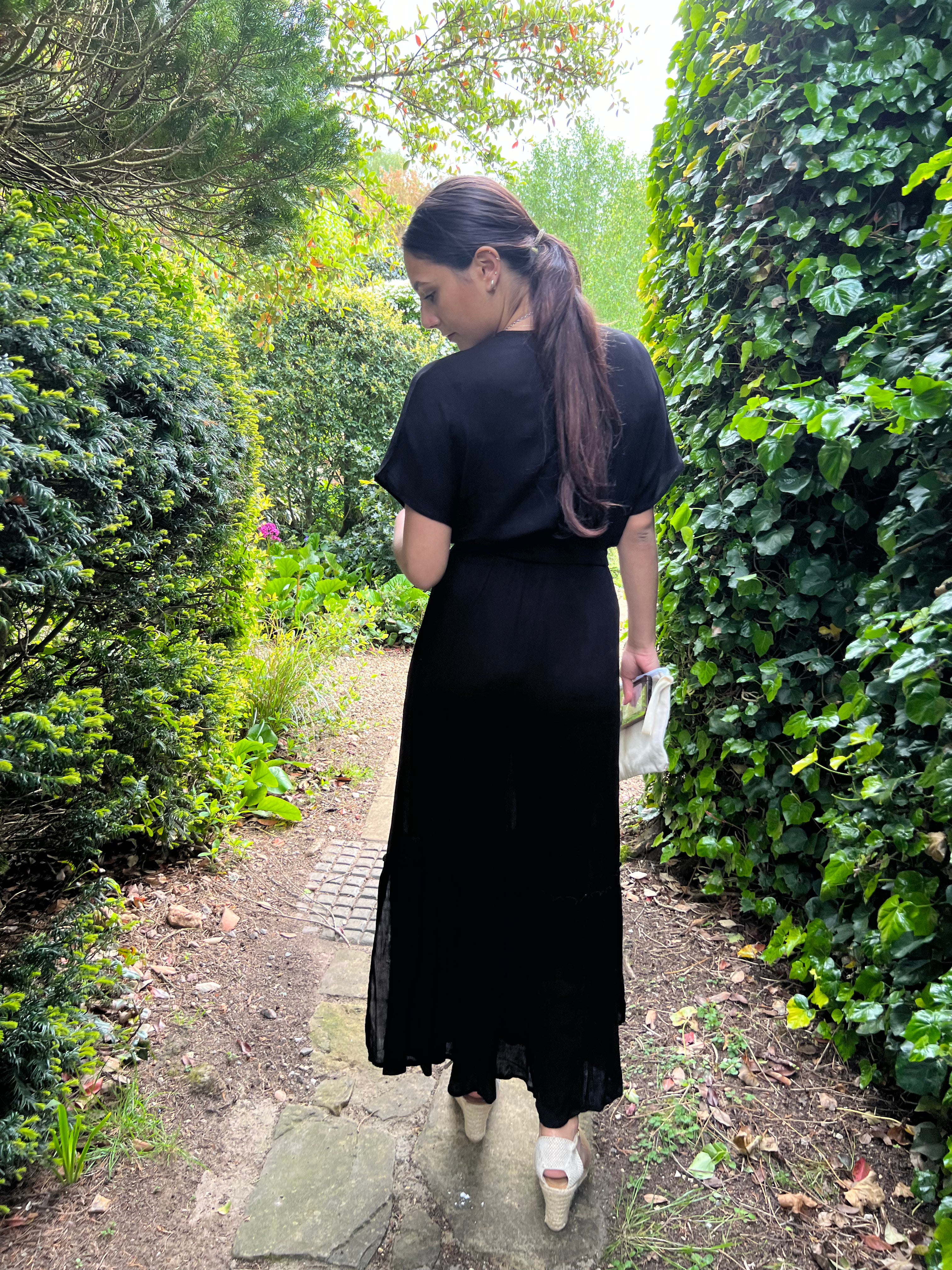 Person standing wearing a talia maxi dress in black by a door outside and trees.