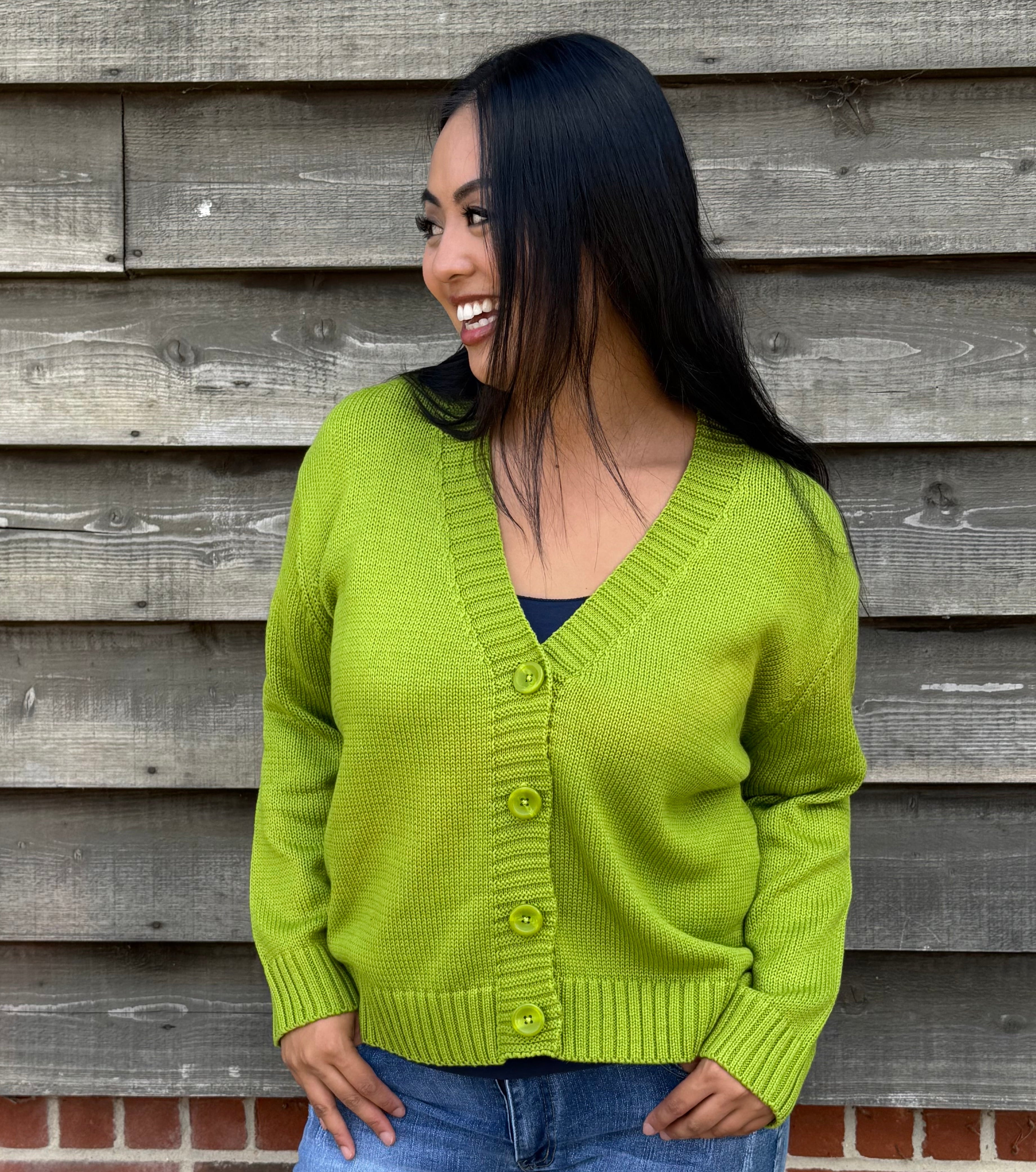 Woman wearing a green cardigan and denim shorts standing against a wooden wall.