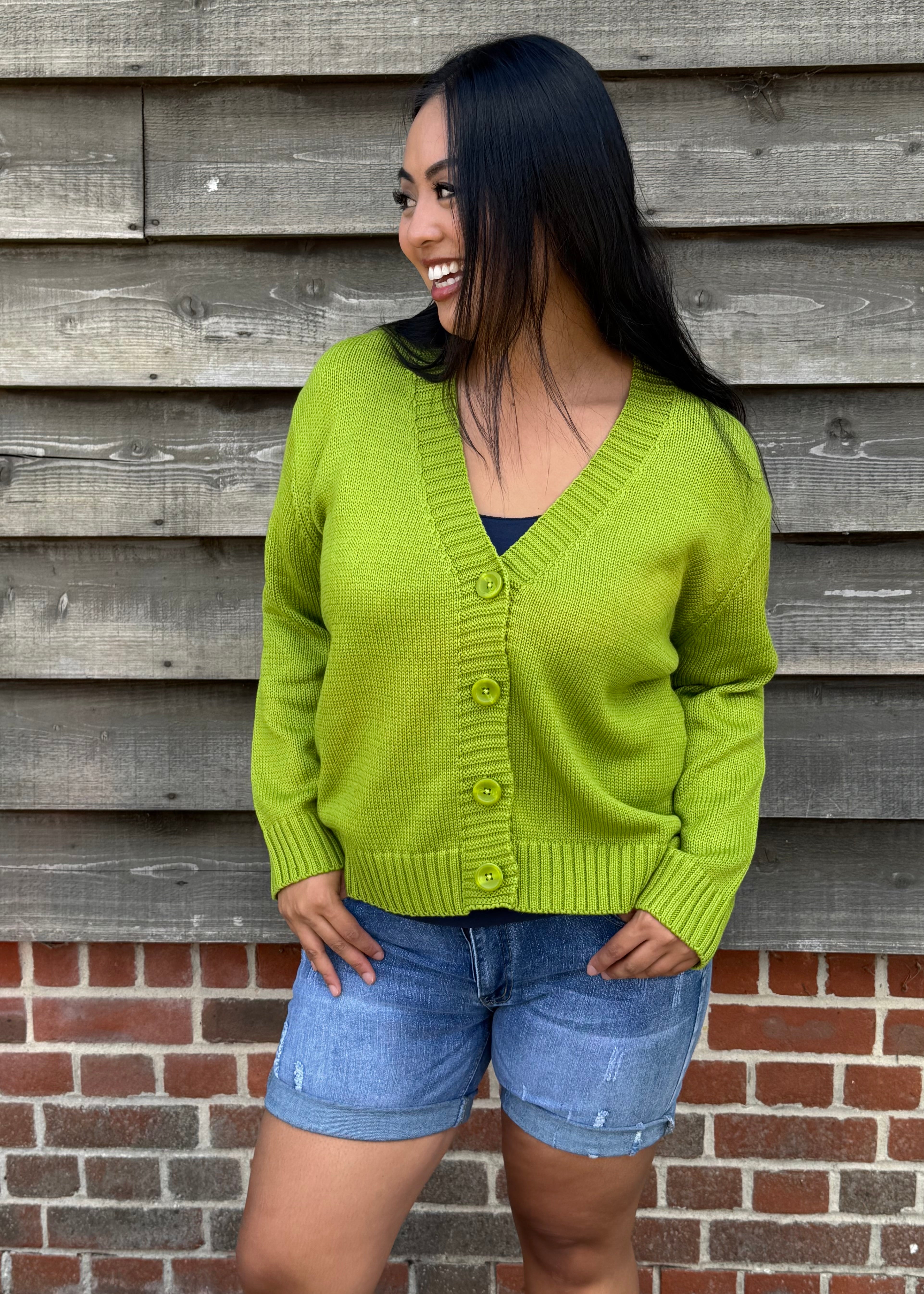 Woman wearing a green cardigan and denim shorts standing against a wooden wall.
