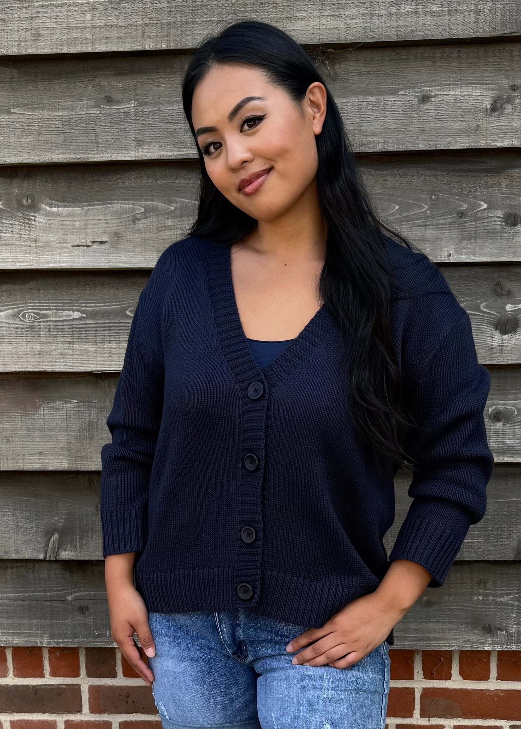 Woman wearing a navy cardigan, denim shorts, and white sandals standing against a wooden wall.
