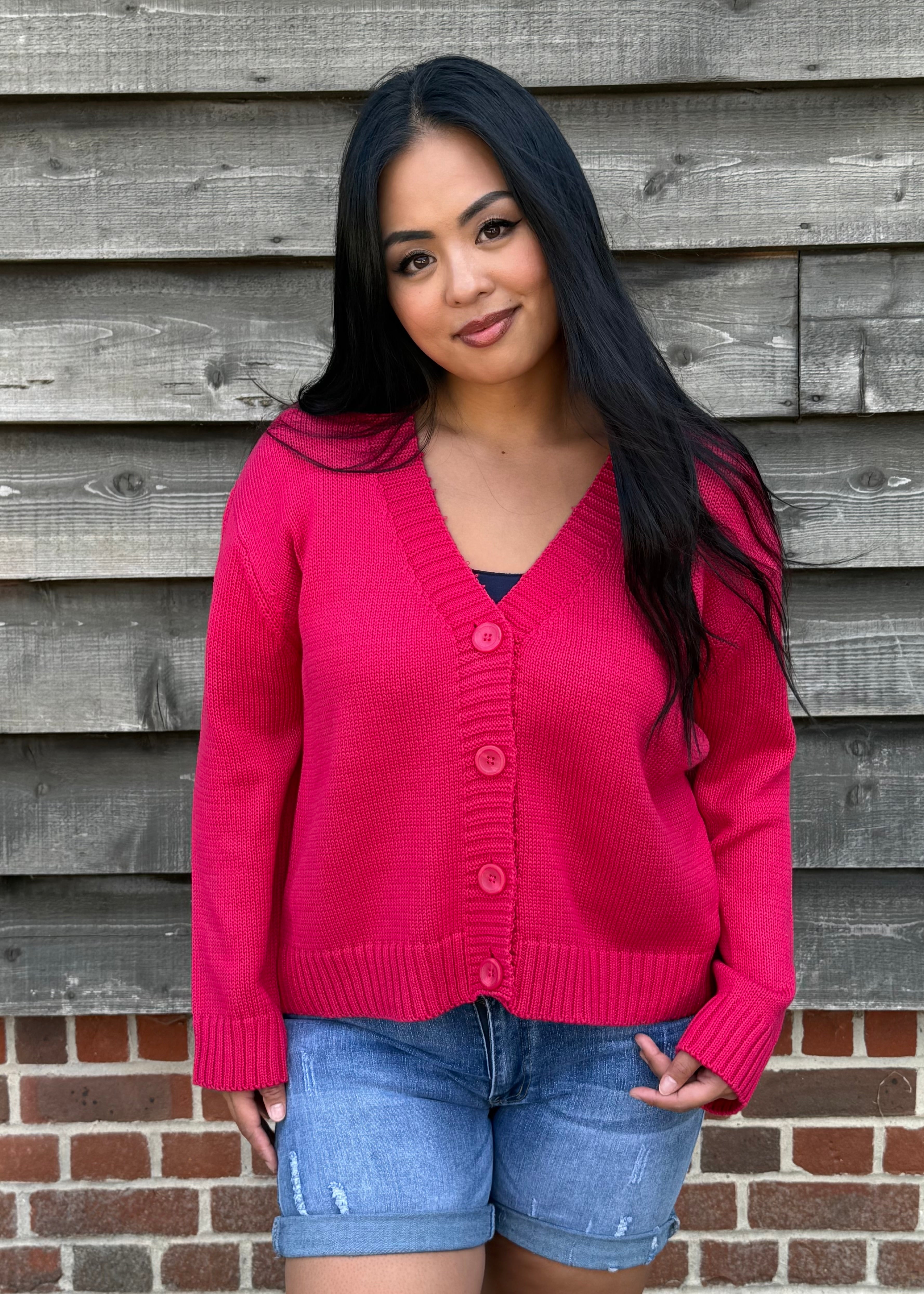 Woman wearing a pink cardigan and denim shorts standing against a wooden wall.