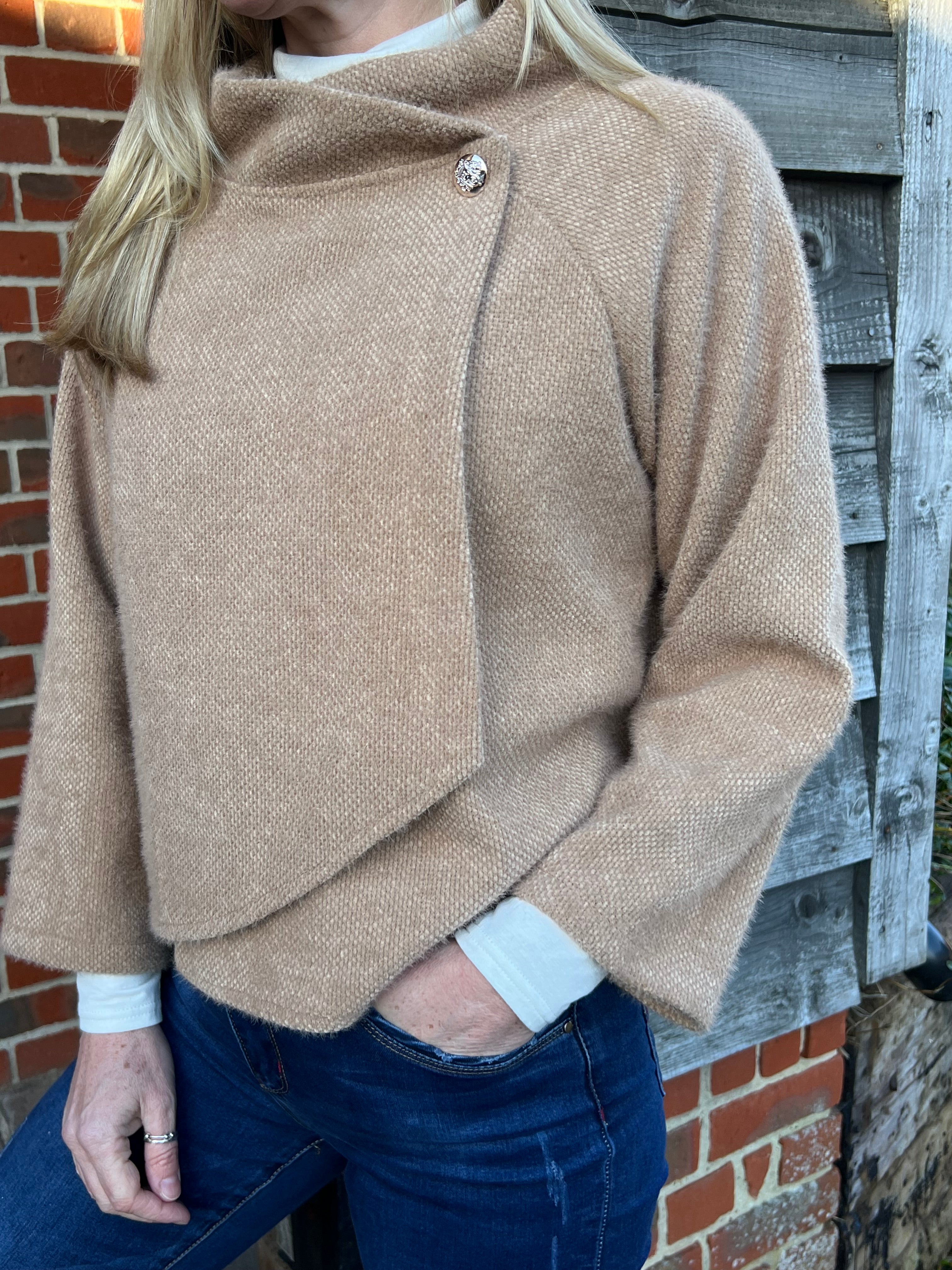 A woman standing outdoors wearing a beige knitted jacket and blue jeans.