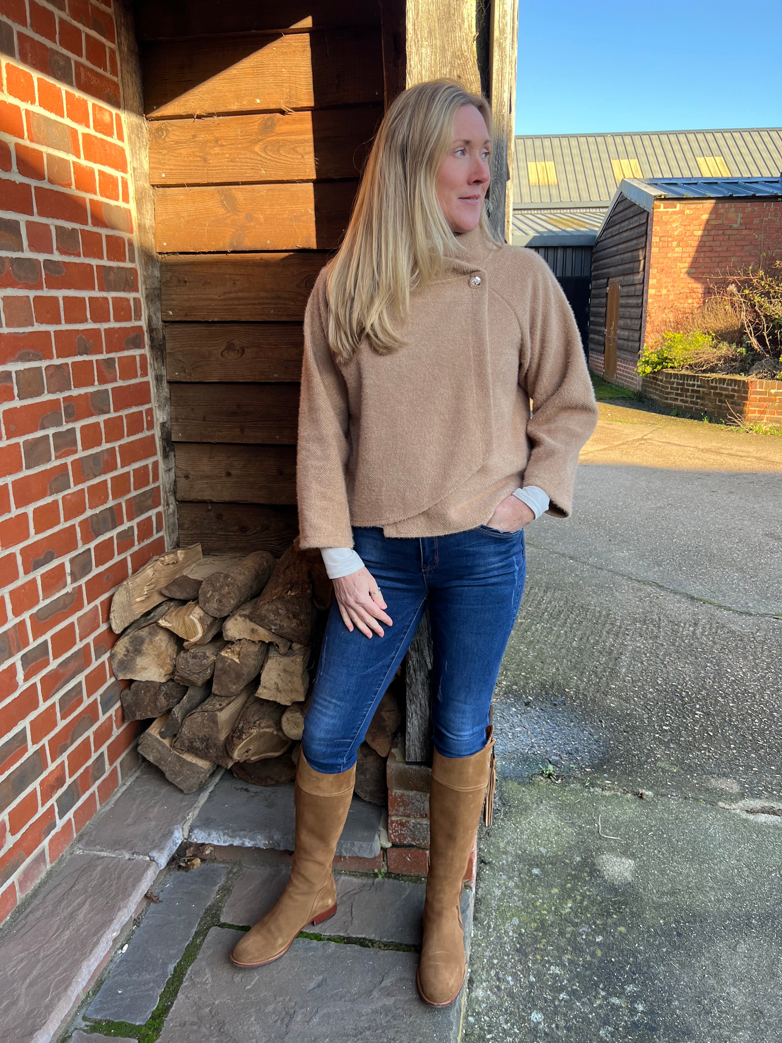 A woman standing outdoors wearing a beige knitted jacket and blue jeans, with brown boots.