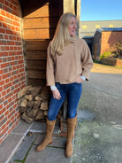 A woman standing outdoors wearing a beige knitted jacket and blue jeans, with brown boots.