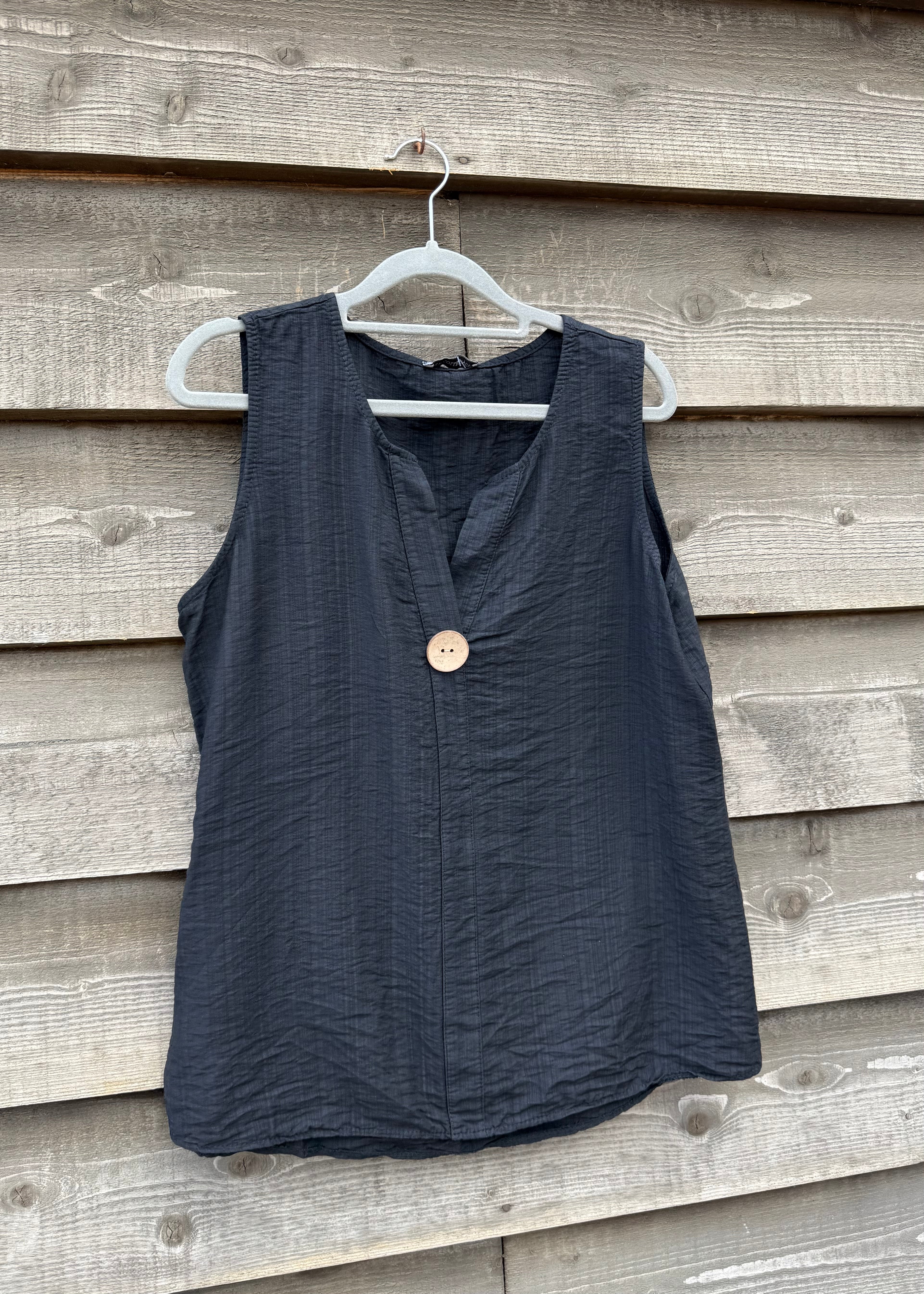 Black sleeveless top with a button on a hanger against a wooden wall.