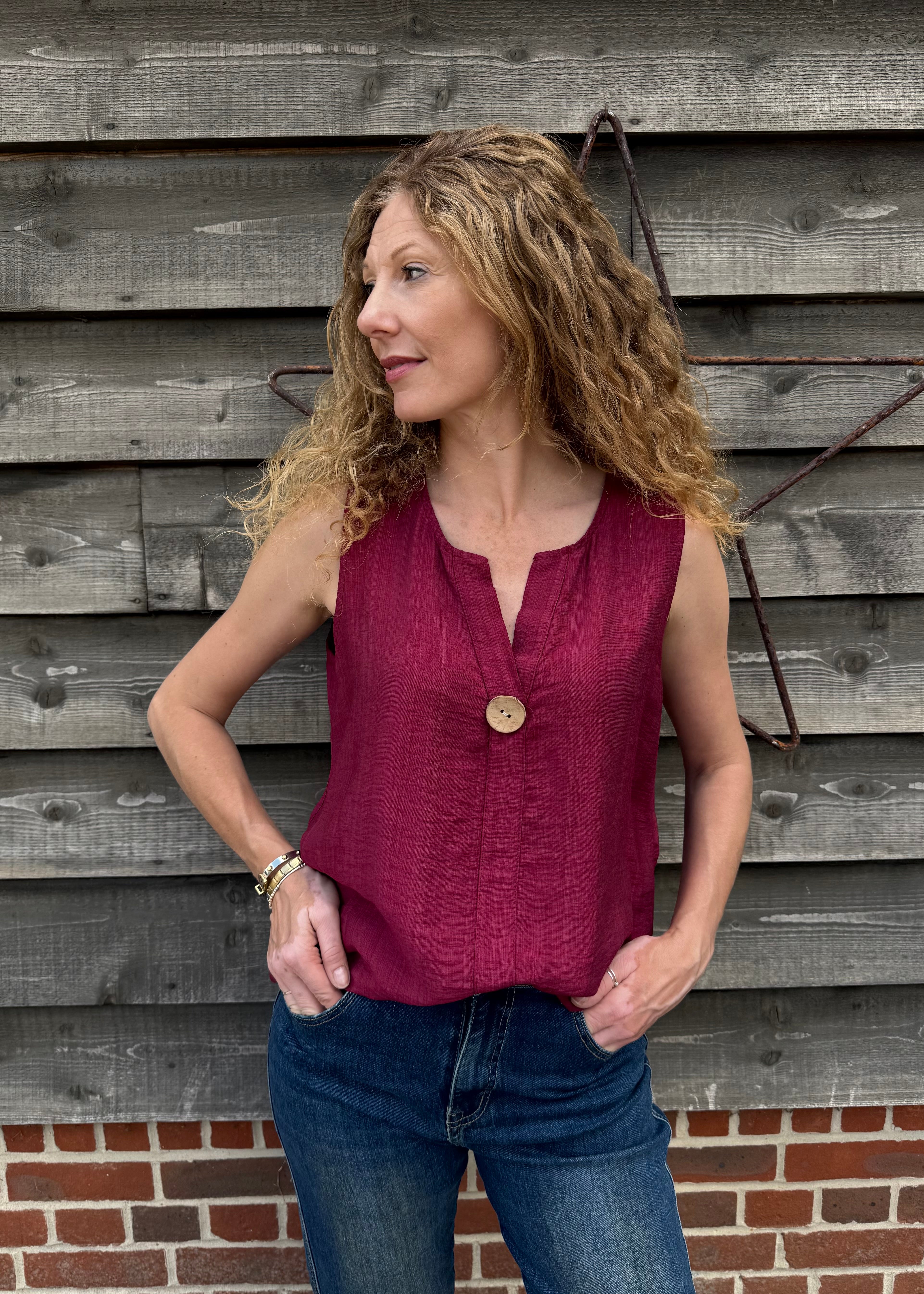 Woman wearing a red sleeveless top and blue jeans standing against a wooden wall.