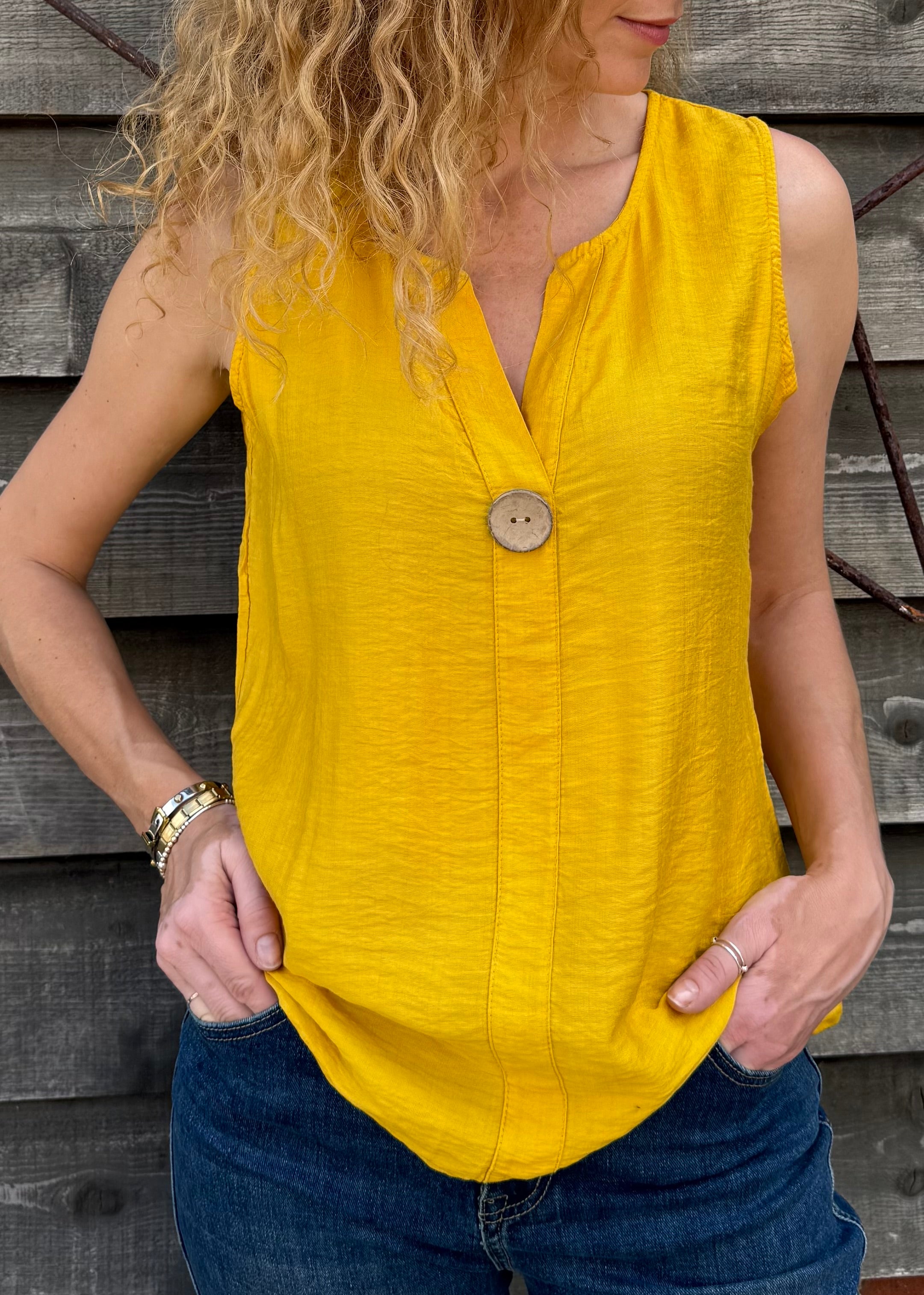 Woman wearing a yellow sleeveless top with a button, standing against a wooden background.