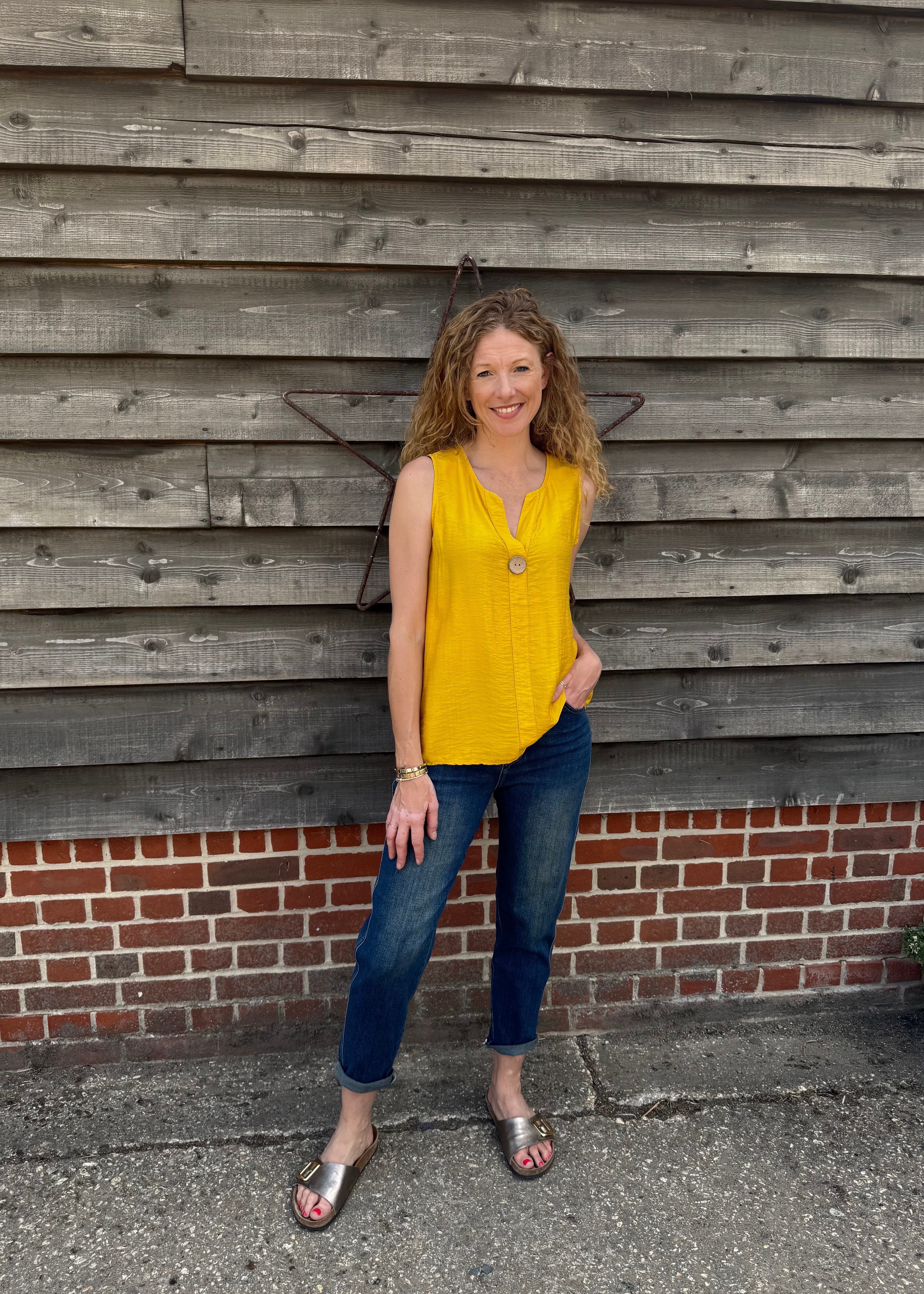 Woman in a yellow top and blue jeans standing against a wooden wall.