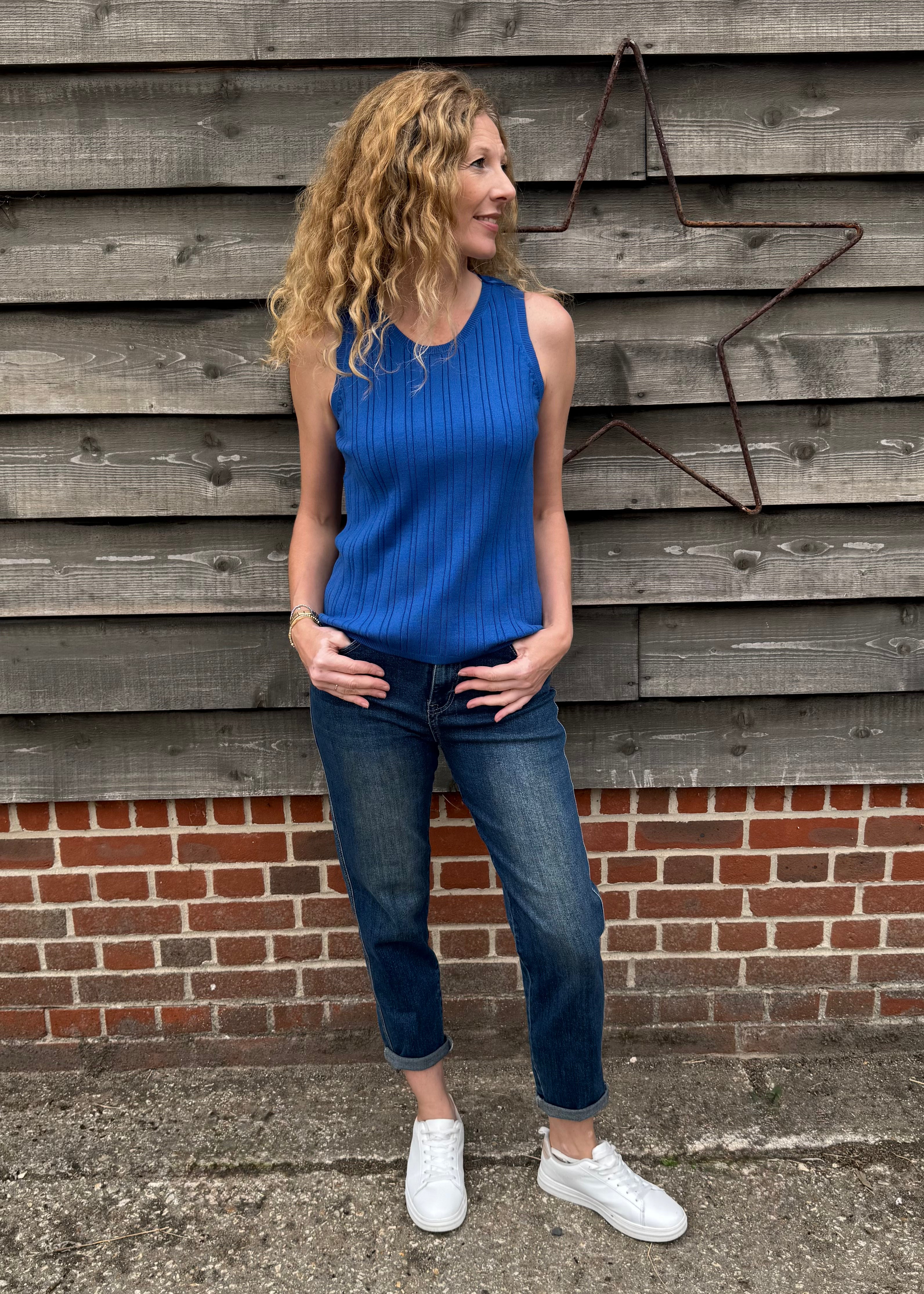 Woman wearing a blue sleeveless top and jeans standing against a wooden and brick wall.