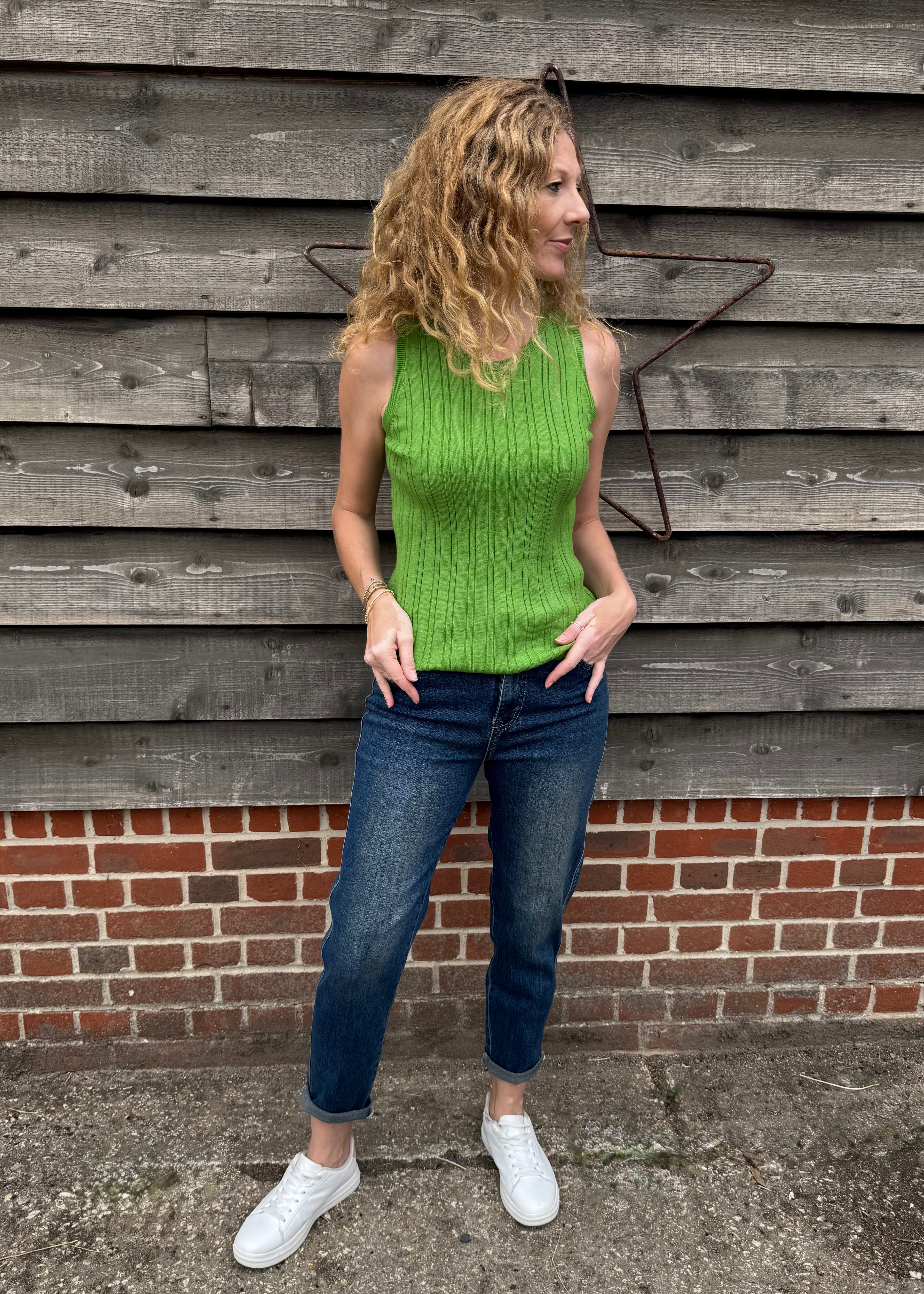 Woman wearing a green sleeveless top and blue jeans standing against a wooden wall.