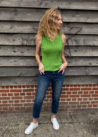 Woman wearing a green sleeveless top and blue jeans standing against a wooden wall.