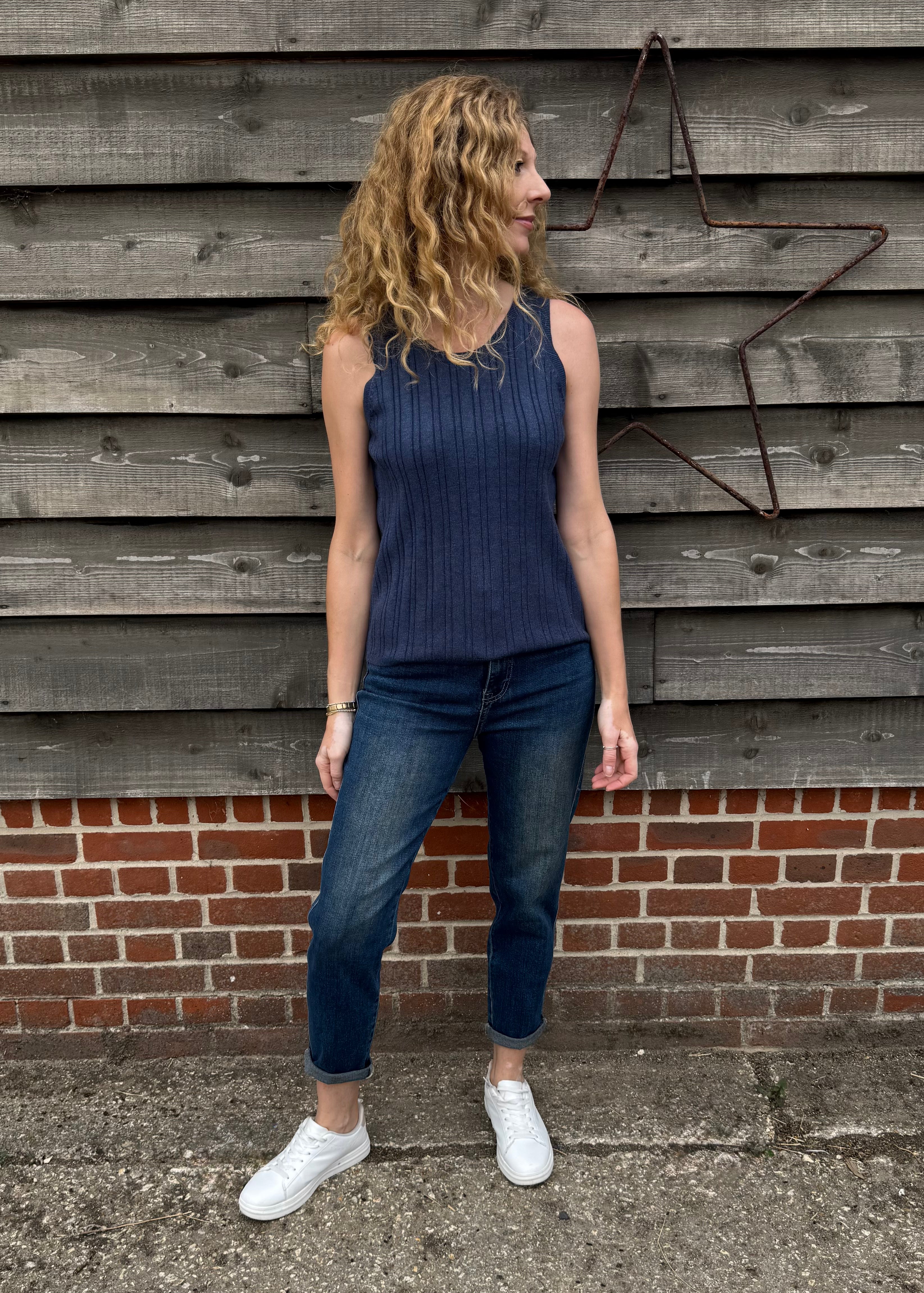 Woman wearing a blue sleeveless top and jeans standing against a wooden wall.
