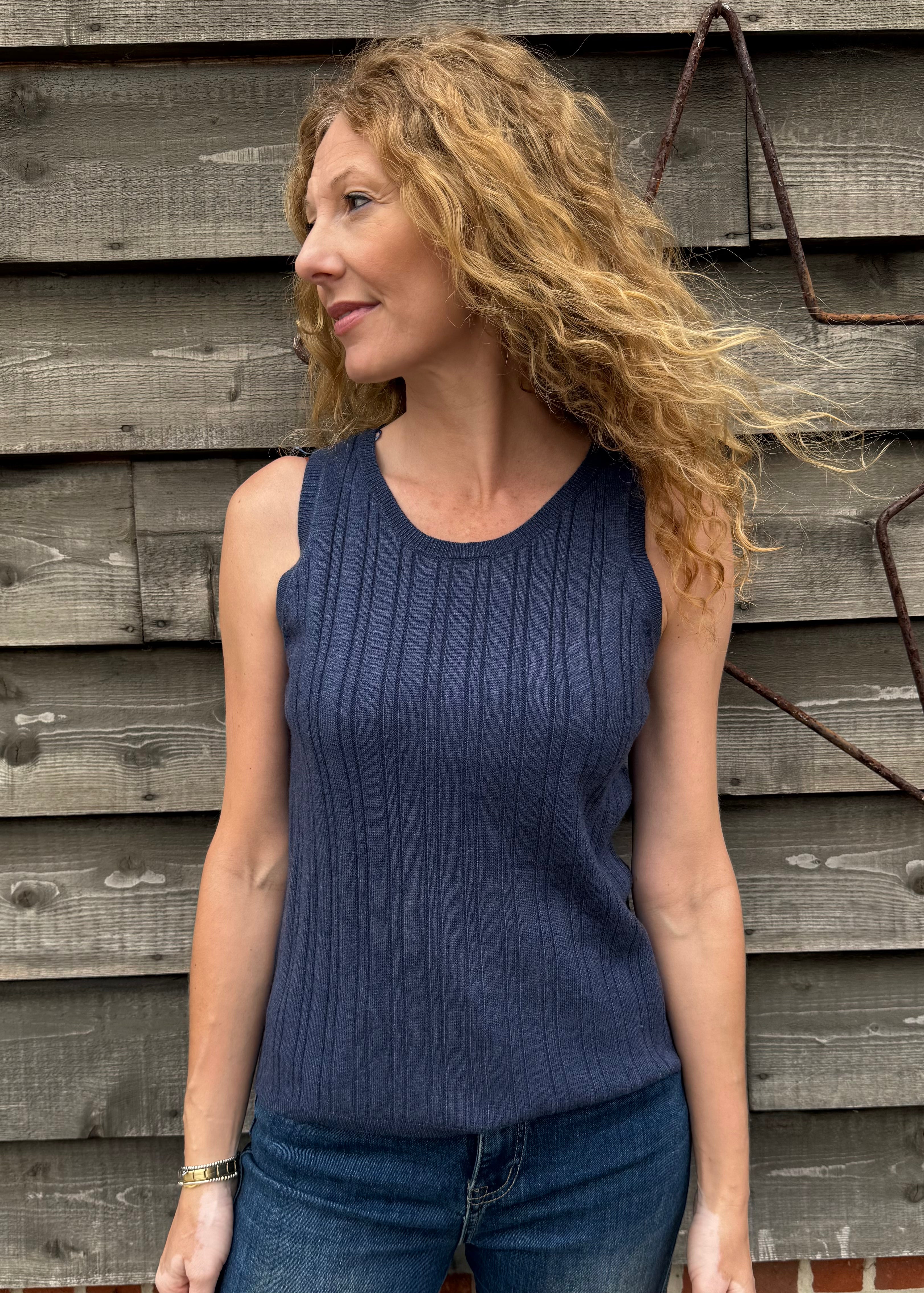 Woman wearing a blue sleeveless top and jeans standing against a wooden wall.