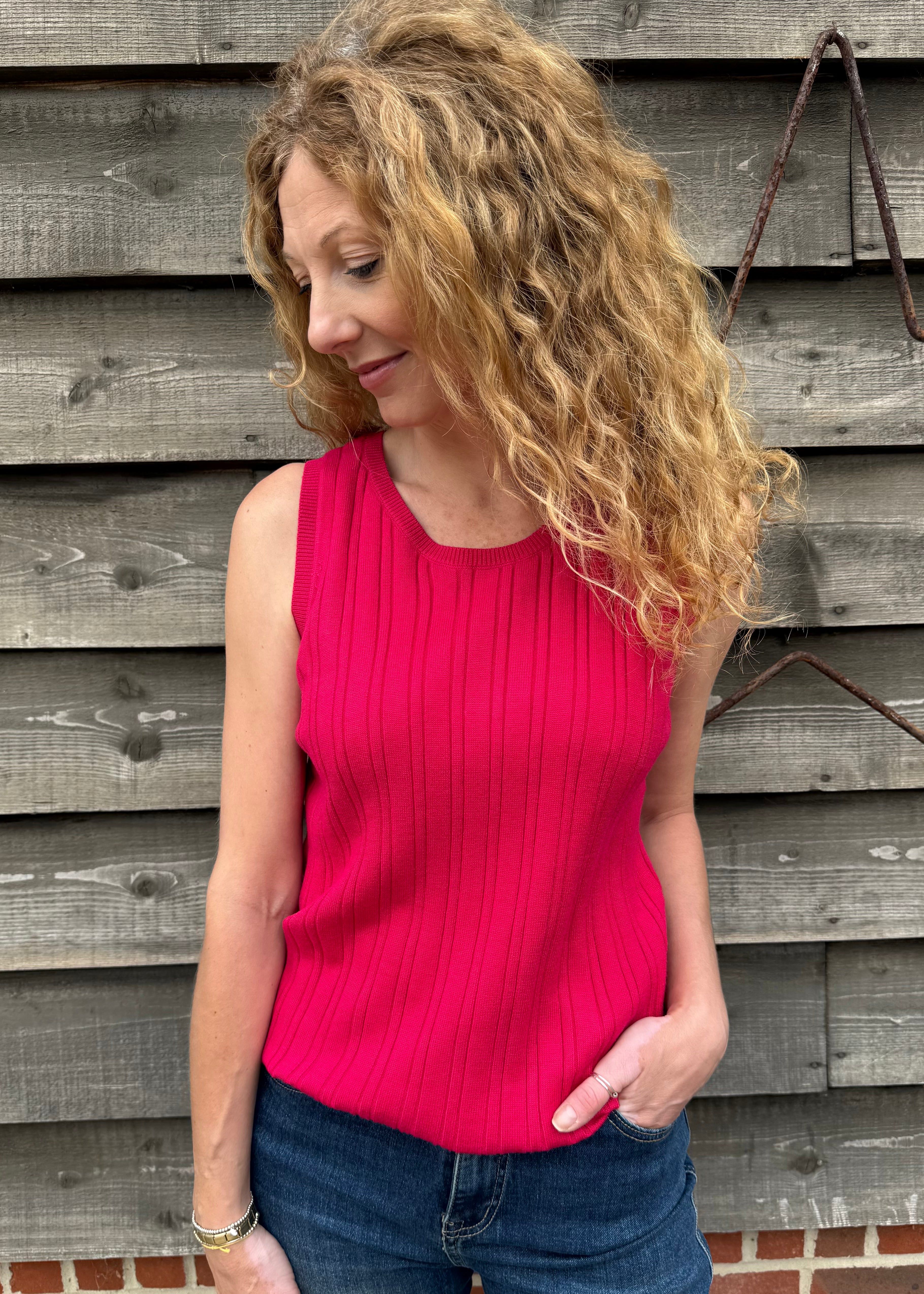Woman wearing a pink sleeveless top and blue jeans standing against a wooden wall.