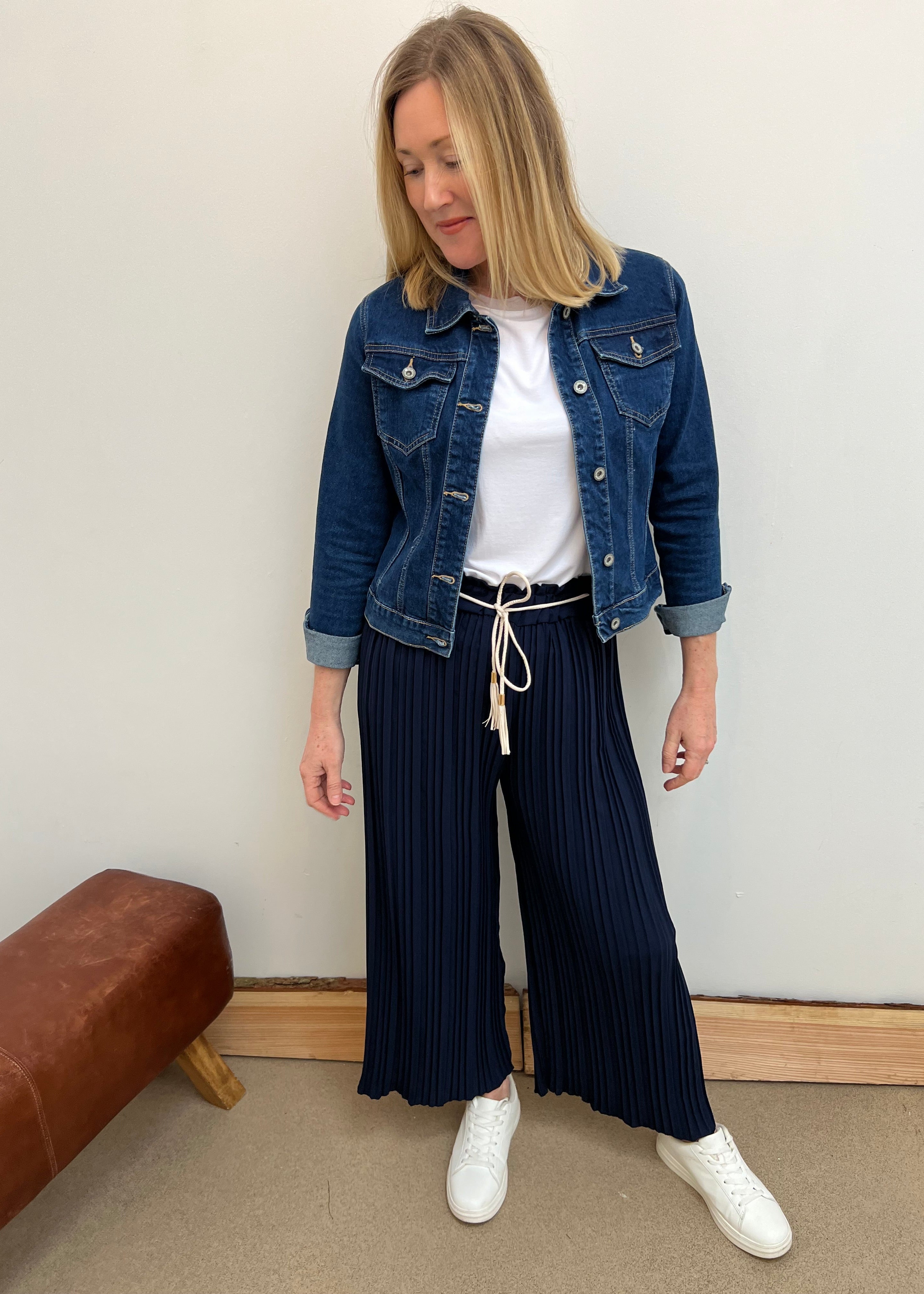 Woman wearing a denim jacket, white shirt, and navy pleated pants against a plain wall.