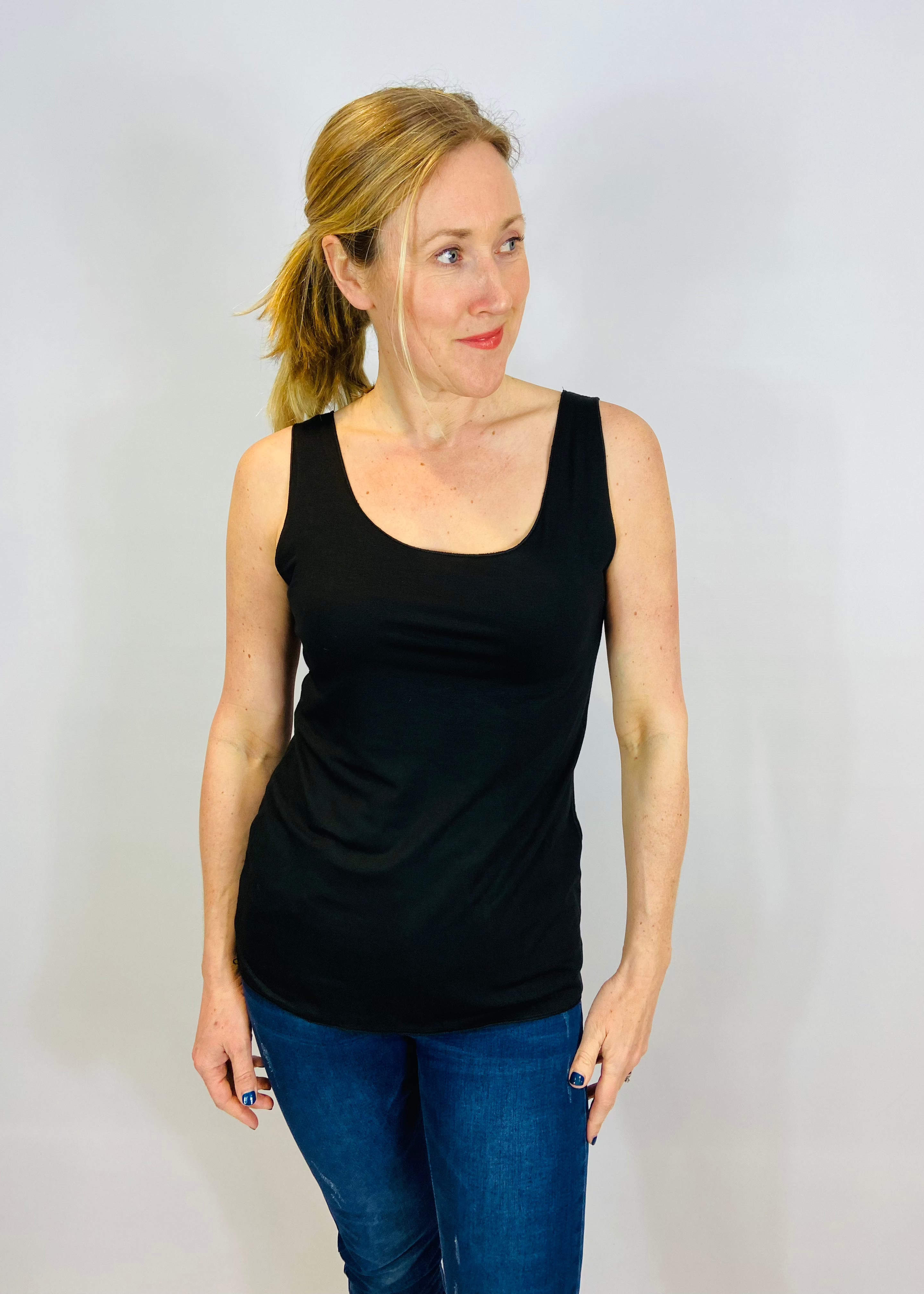 A woman wearing a black sleeveless tank top and blue jeans standing against a plain background.