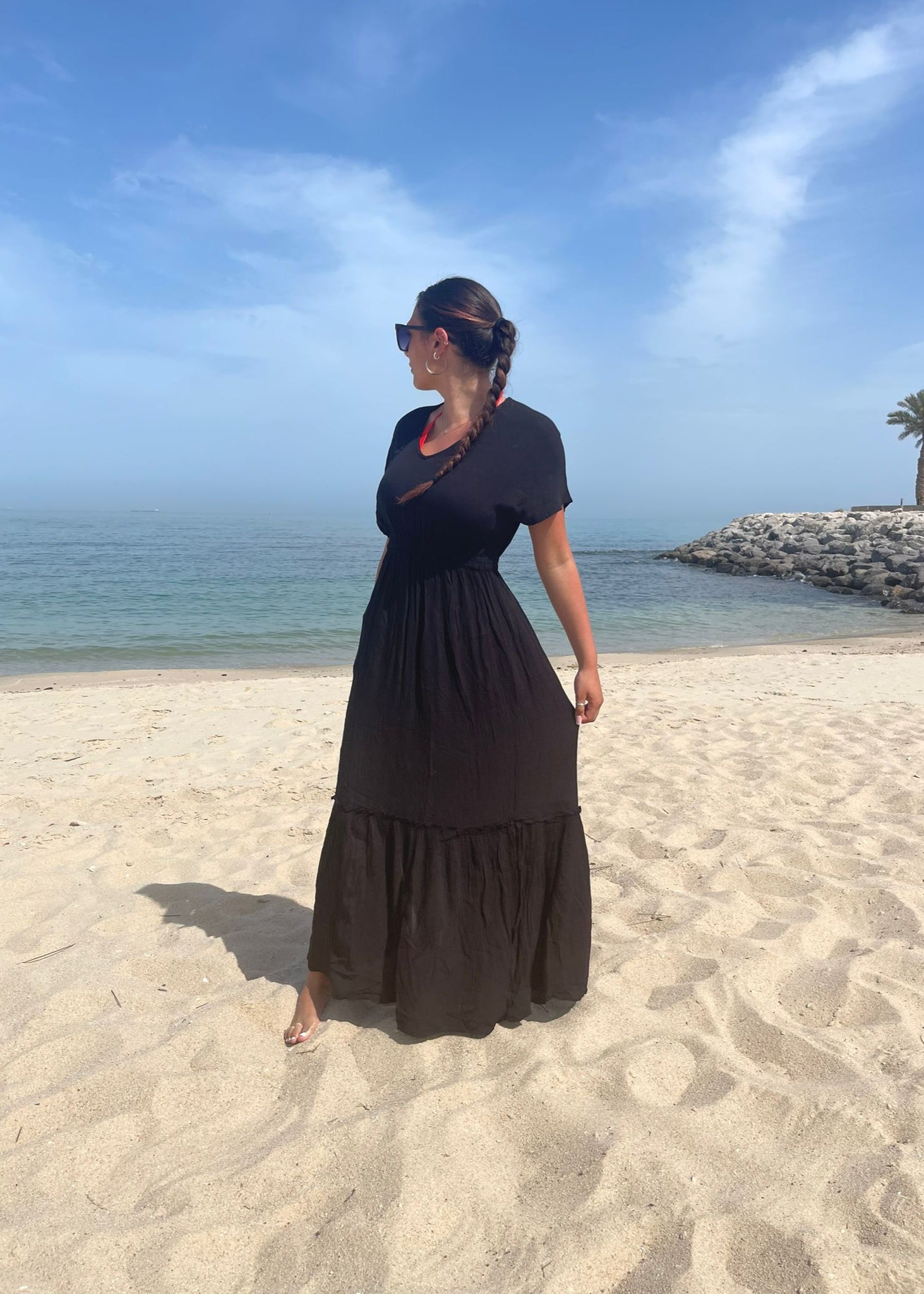 A person standing on a sandy beach wearing a navy maxi dress with short sleeves and a knee-length skirt, with a clear blue sky and some palm trees in the background.