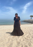 A person standing on a sandy beach wearing a navy maxi dress with short sleeves and a knee-length skirt, with a clear blue sky and some palm trees in the background.